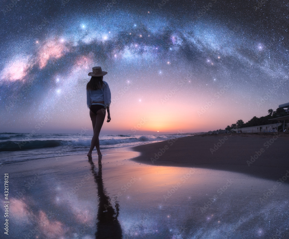 拱形银河和戴着帽子的年轻女子在沙滩上，星空映照在近处的水中