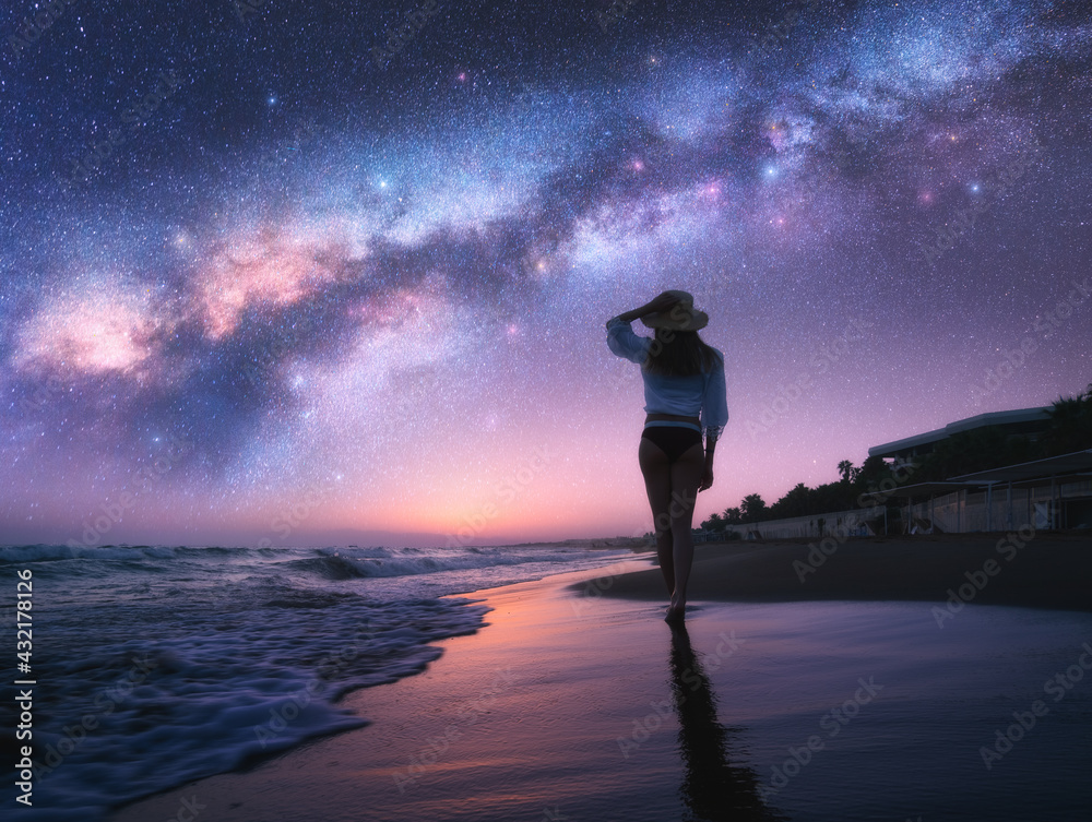 银河系和戴帽子的年轻女子在沙滩上，紫色的星空映照在近处的水中