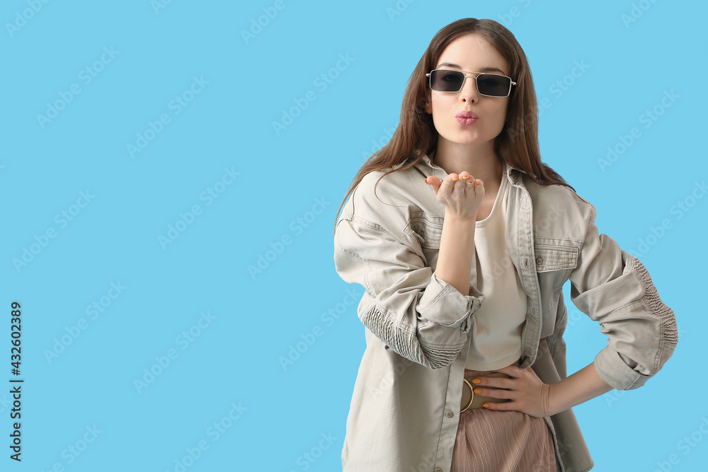 Beautiful young woman with stylish sunglasses blowing kiss on color background