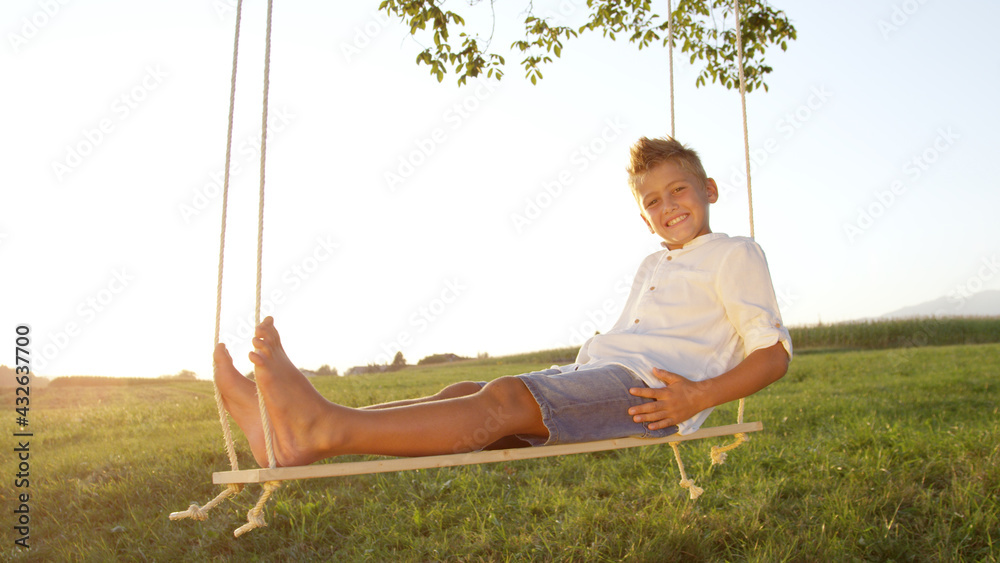 CLOSE UP Young boy relaxing on a wooden swing at sunset, posing for the camera.