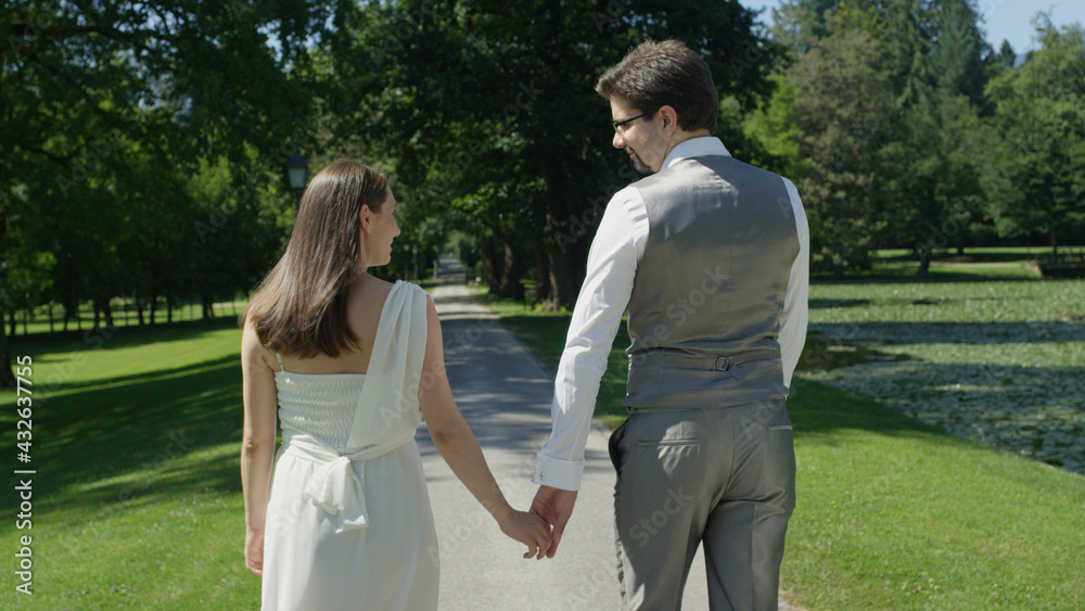 BACK VIEW: Young happy couple holding hands gaze at each other walking in park