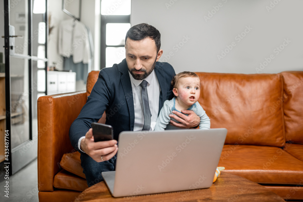年轻的父亲在家里用笔记本电脑工作，一边看电话，一边照顾他的小儿子。多
