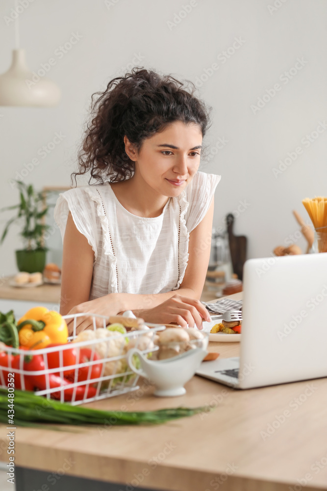 年轻女子在厨房吃饭时使用笔记本电脑