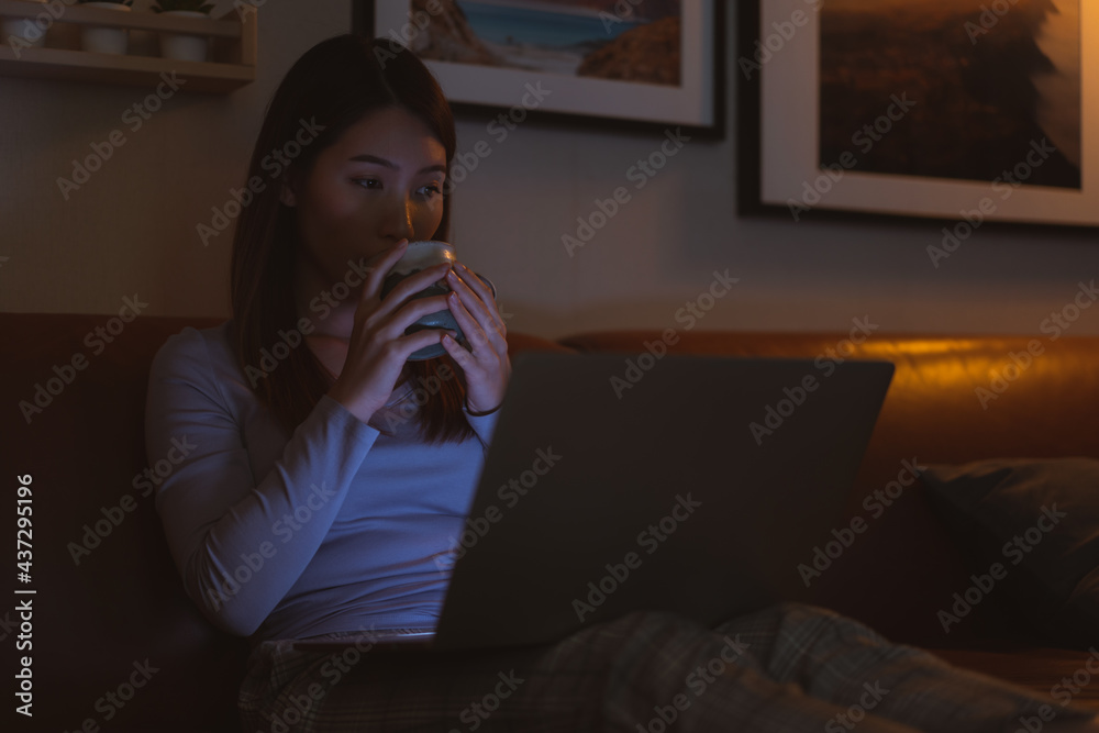 亚洲女子晚上拿着热牛奶杯在笔记本电脑上看网络电影