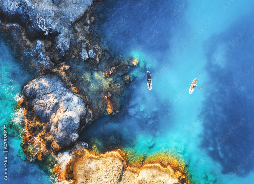 夏日日落时，人们在蓝色海洋、岩石和树木上的漂浮支撑板上的鸟瞰图。蓝色滞后