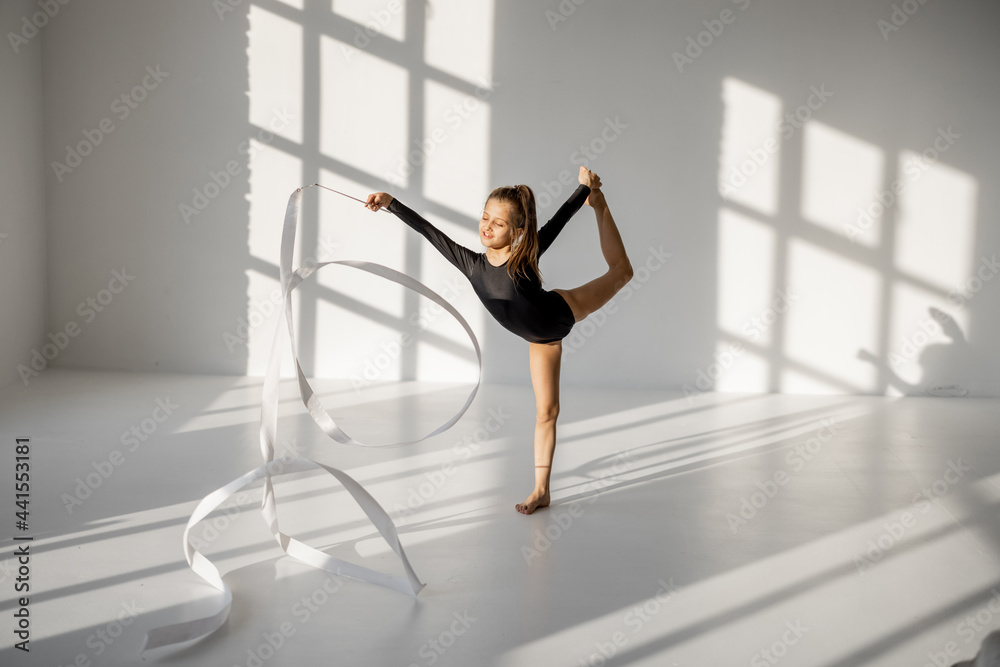 Little girl practising rhythmic gymnastics with a gymnastic tape at white sunny dance room. Wide vie