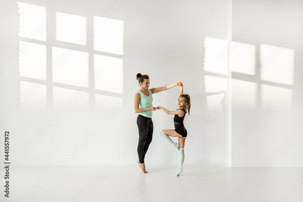 年轻的妈妈或体育教练在阳光明媚的体育教室里训练小女孩进行艺术体操。