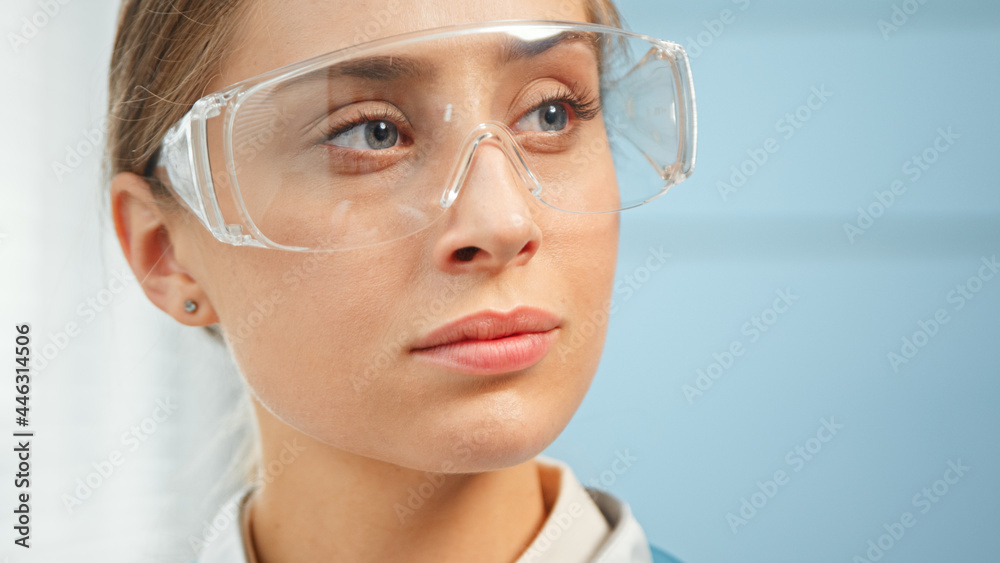 Exhausted young woman medical nurse in protective glasses looks straight standing in hospital room