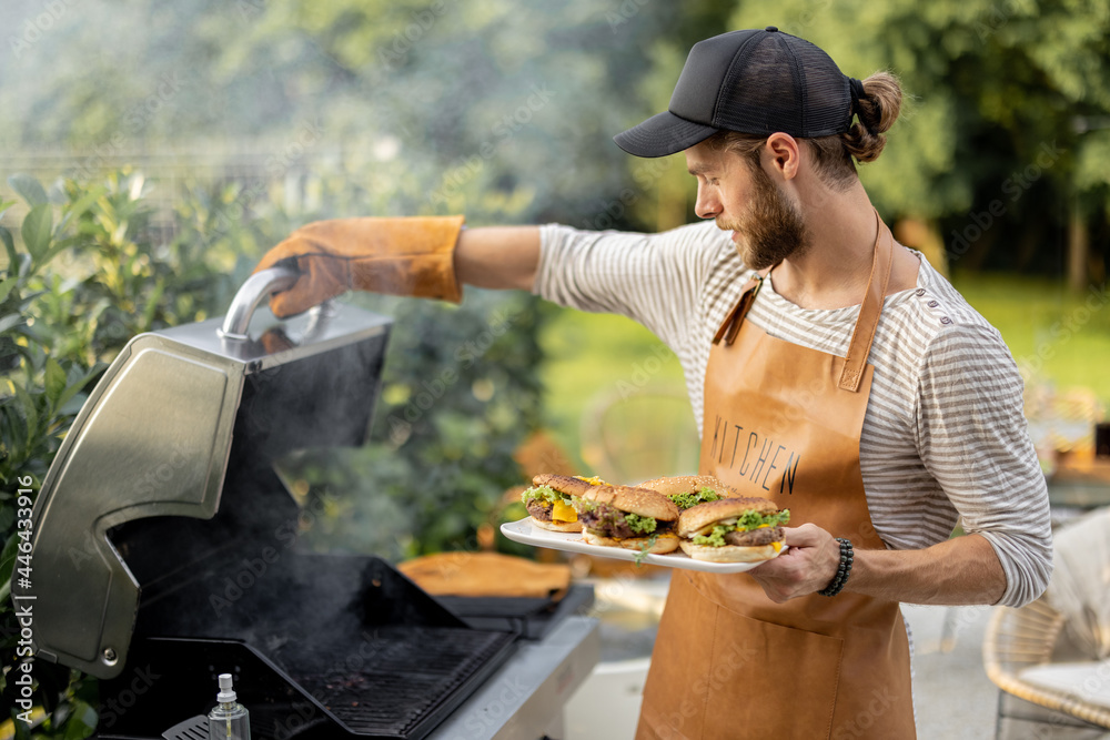 戴着帽子和围裙的英俊男子在后院的烤架上做汉堡。户外烹饪和美式烹饪