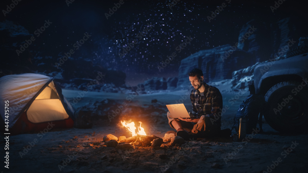 峡谷露营夜帐篷：男旅行者坐在篝火旁用笔记本电脑。数字男人