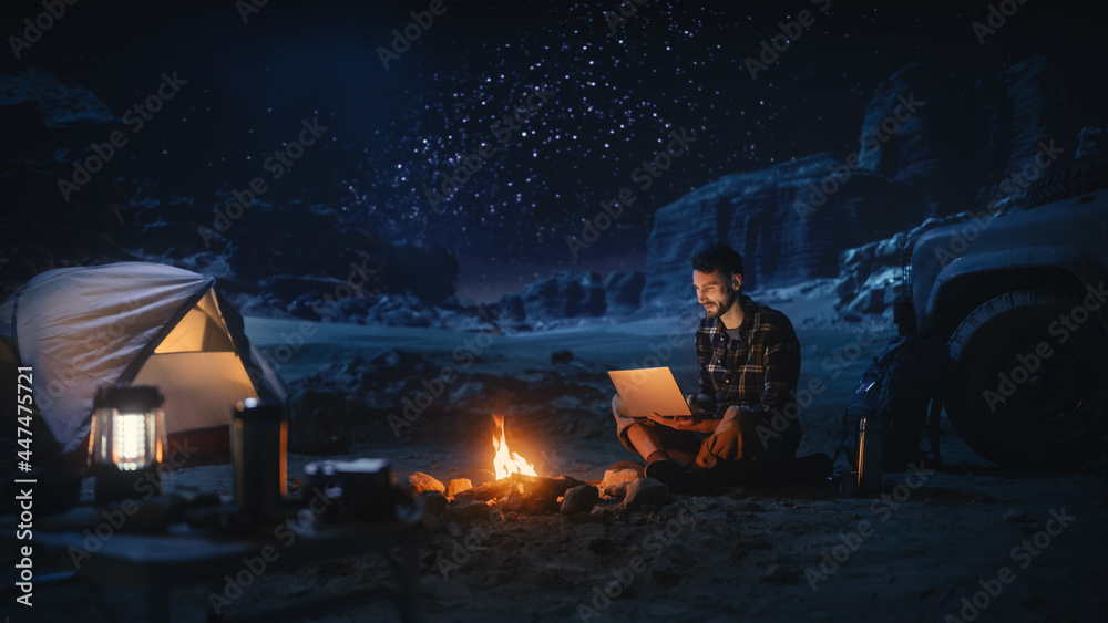 坐在篝火旁的男性旅行者在峡谷露营时使用笔记本电脑。男子做D