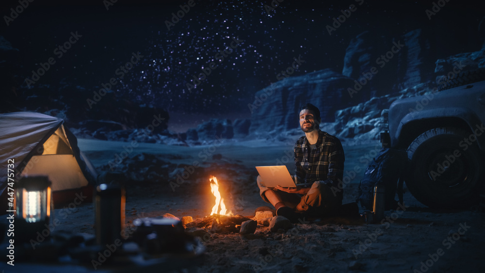 峡谷露营夜帐篷：男旅行者坐在篝火旁用笔记本电脑。男人在数码上