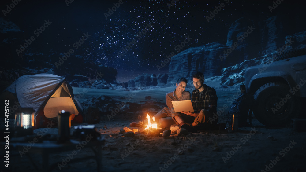 快乐情侣夜间在峡谷自然露营，使用笔记本电脑，坐在篝火旁。两个Tr