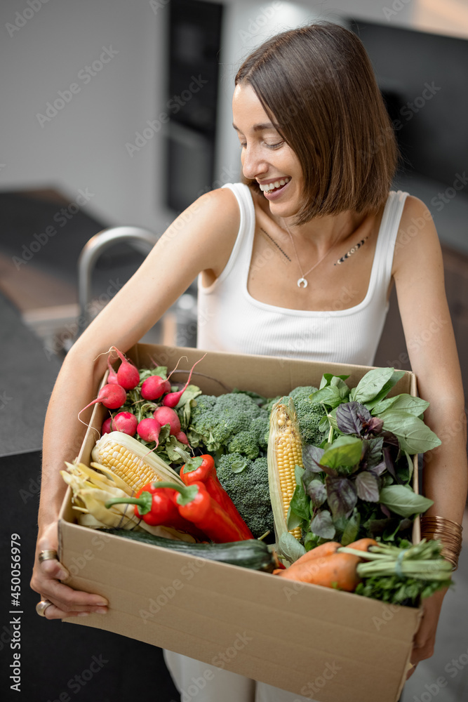 Young woman with fresh vegetables packed in cardboard box at kitchen. Delivering and buying food onl
