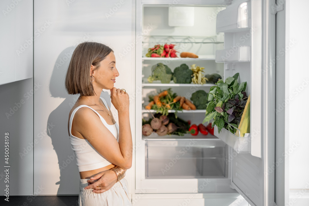 年轻女子站在现代厨房里装满新鲜蔬菜的冰箱旁。健康的素食主义者