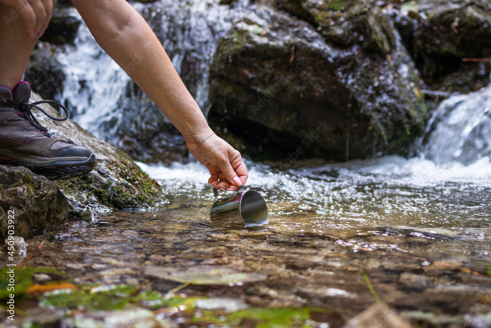 Thirsty hiker scoops fresh water into cup from stream in mountains. Refreshment during hiking in nat