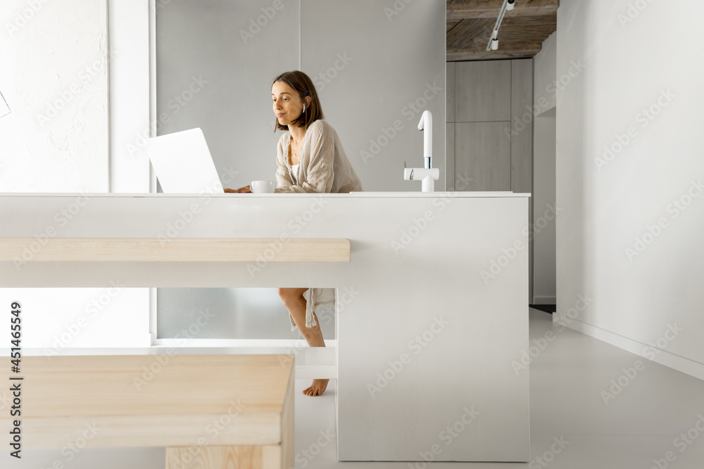一位年轻女士站在现代明亮的一室公寓的餐桌旁，用笔记本电脑工作。