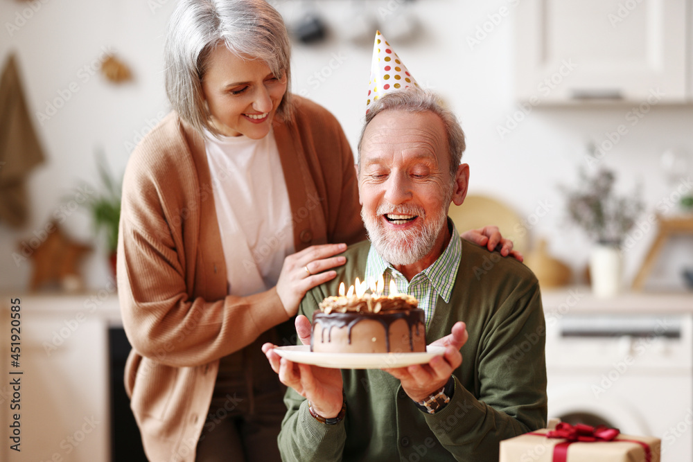 生日庆祝活动中，戴着派对帽子的快乐老人手持点燃蜡烛的巧克力蛋糕