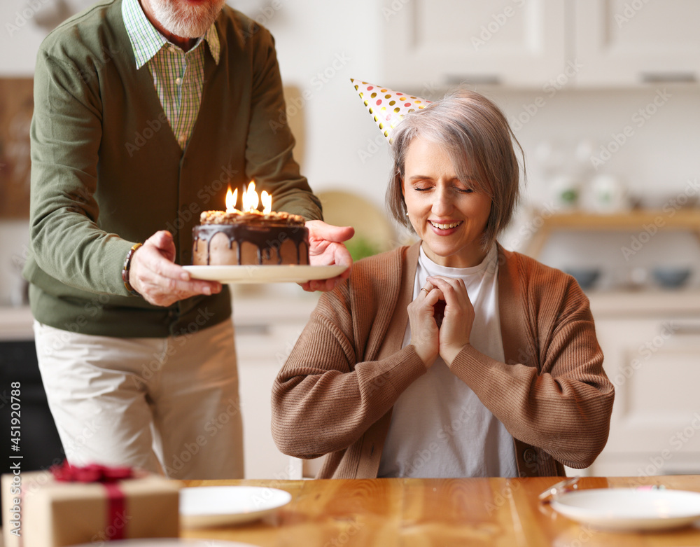 Happy elderly woman in party hat closing her eyes, she makes a wish, while her husband holds a choco
