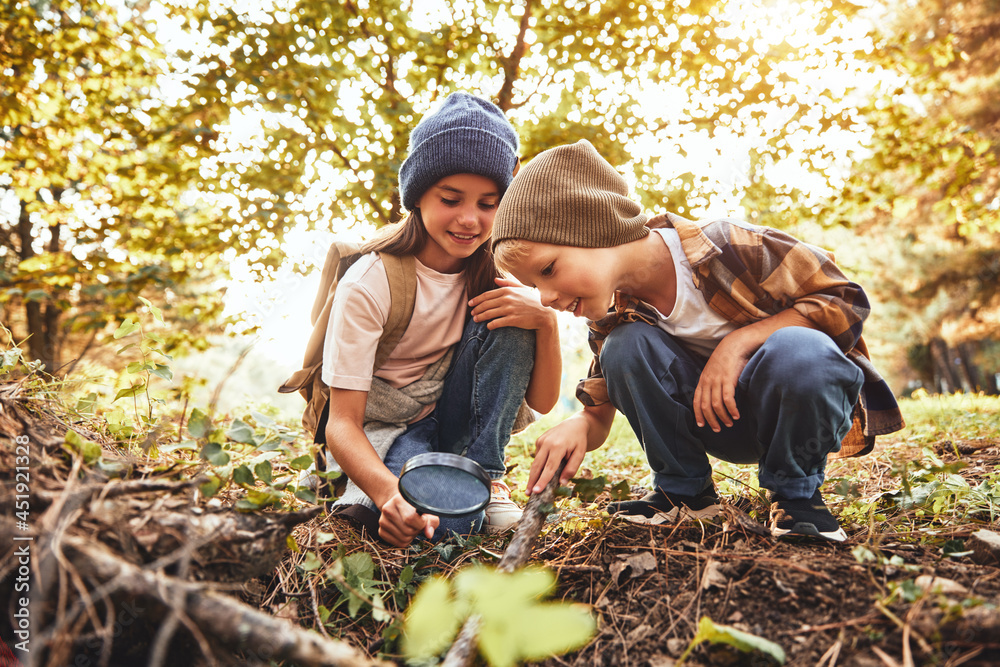 Two little kids in warm hats with backpacks examining tree bark through magnifying glass in forest