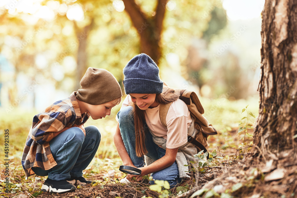 两个戴着温暖帽子、背着背包的小孩在森林里用放大镜检查树皮