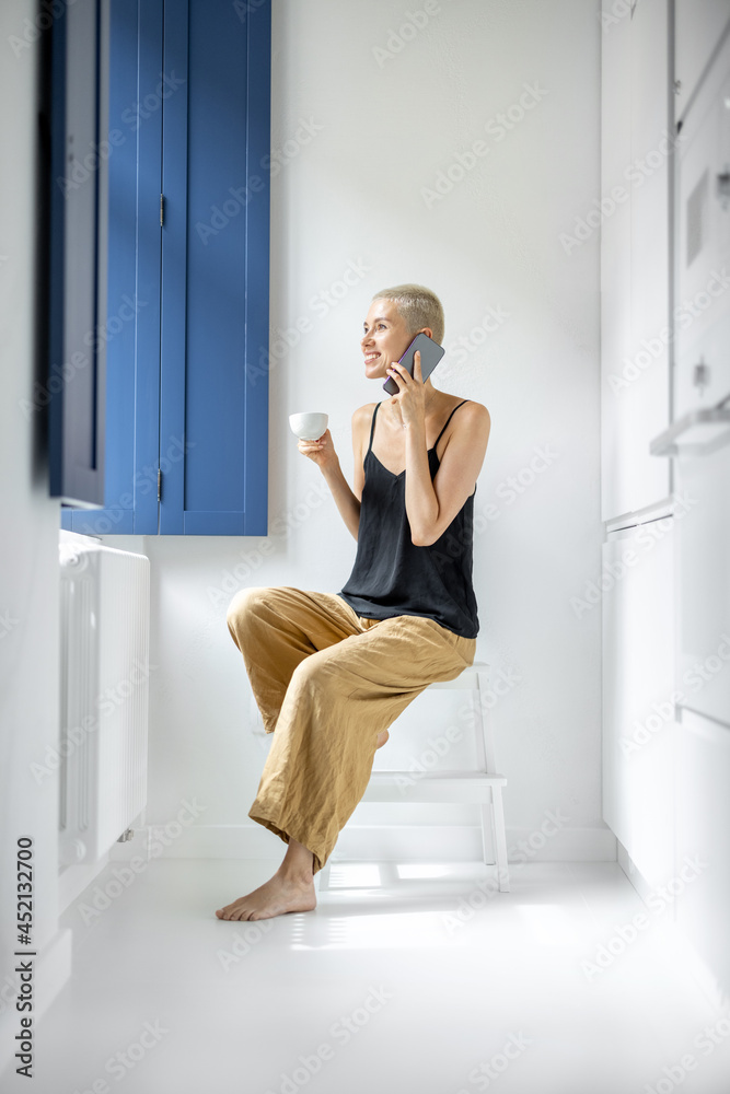 年轻时尚的女人坐在现代和白色房子的窗户附近，拿着电话和咖啡