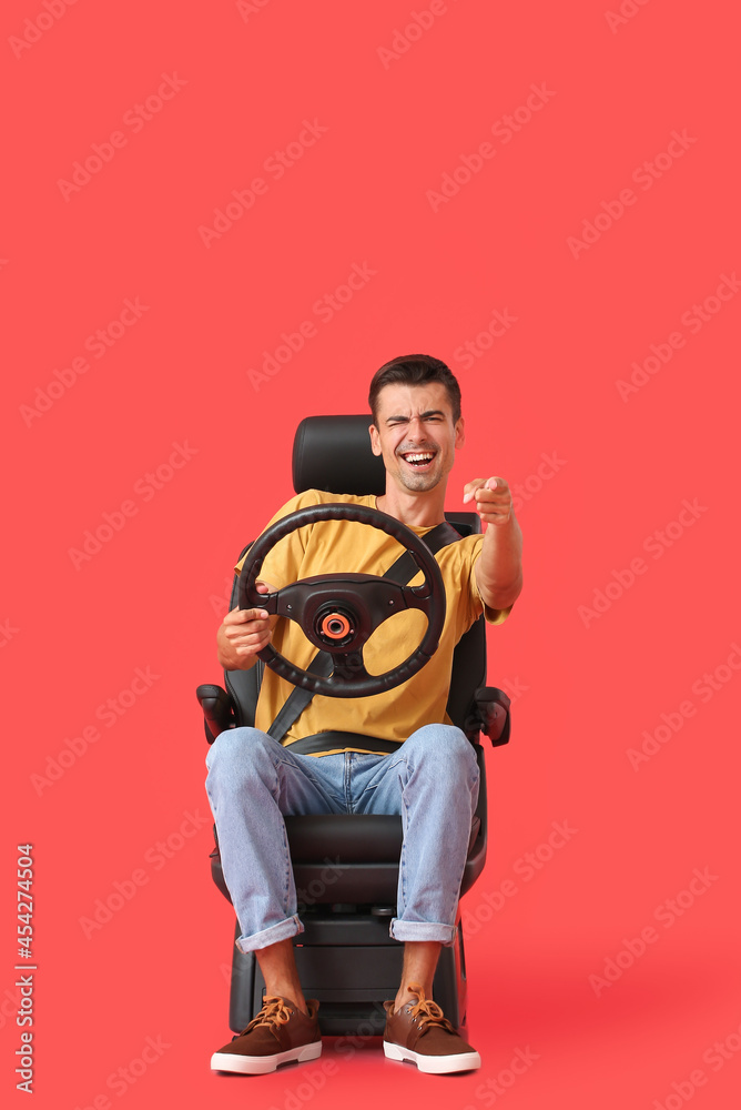 Emotional man in car seat and with steering wheel pointing at viewer on color background