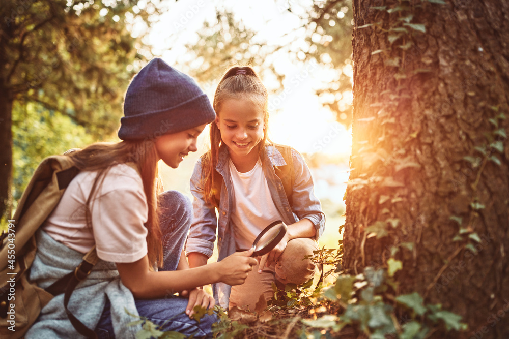 Two little girls in warm hats with backpacks examining tree bark through magnifying glass in forest