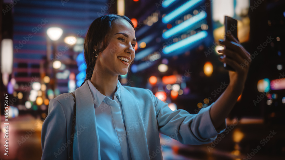 美丽的年轻女子在霓虹灯下的夜城街道上使用智能手机进行视频通话。Portrai