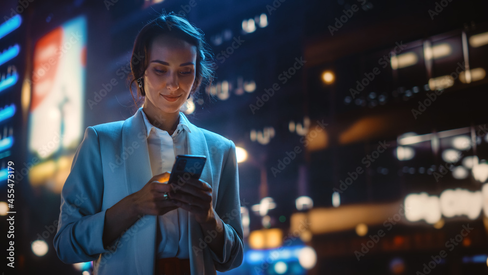 漂亮的年轻女人用智能手机穿过充满霓虹灯的夜城街道。波特拉伊