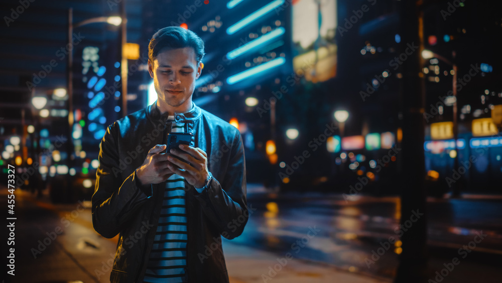 男人用智能手机穿过充满霓虹灯的夜城街道。微笑时尚的男人用