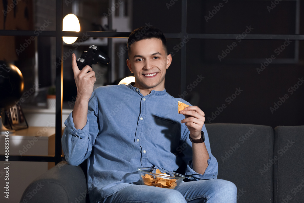 Handsome young man eating tasty nachos and playing video game at home late in evening