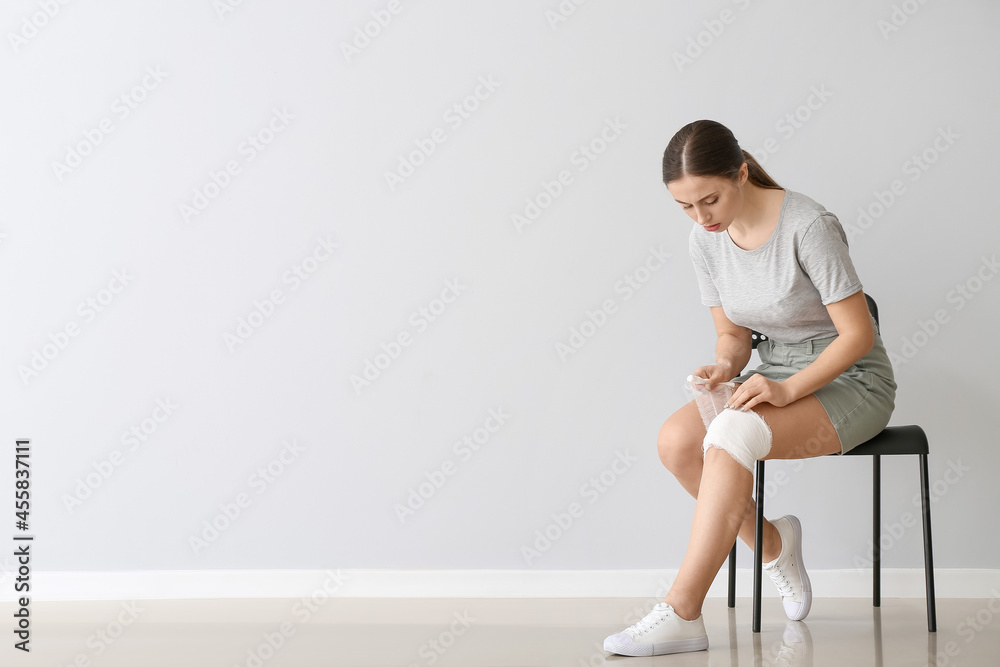 Young woman applying bandage onto her knee near light wall