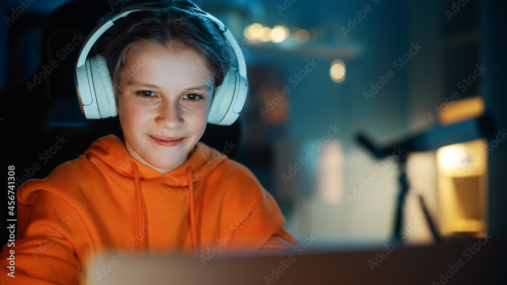 戴着耳机的聪明男孩在家里舒适的暗室里使用笔记本电脑。快乐的青少年Browsi