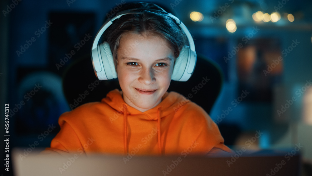 戴着耳机的聪明男孩在家里舒适的暗室里使用笔记本电脑。快乐的青少年Browsi