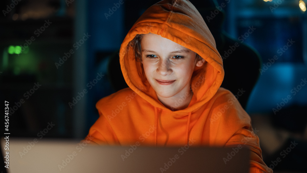 Cute Young Boy in a Hoodie Using Laptop Computer in Cozy Dark Room at Home. Happy Teenager Browsing 