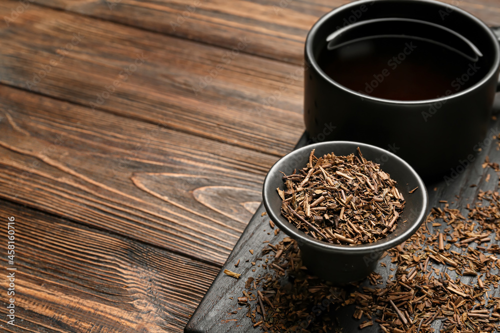 Bowl with dry hojicha green tea and cup of hot beverage on wooden background