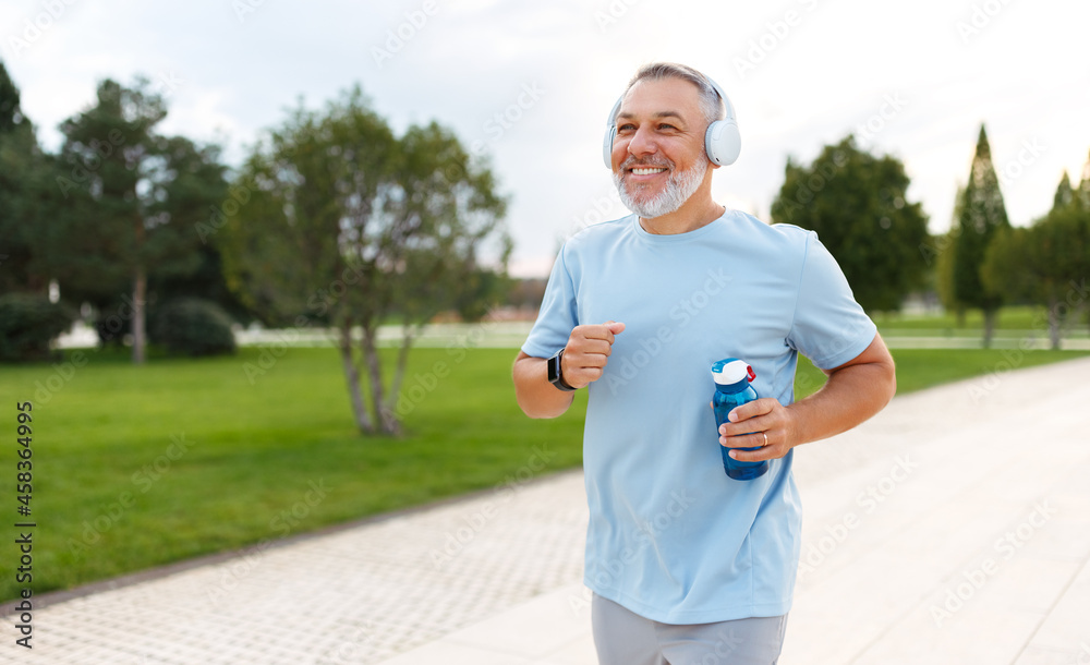 快乐的成熟男人戴着耳机，手里拿着水瓶在城市公园外慢跑
