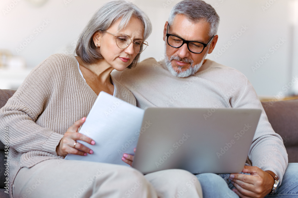 一对忧心忡忡的资深家庭夫妇坐在家里的沙发上用笔记本电脑在线支付账单