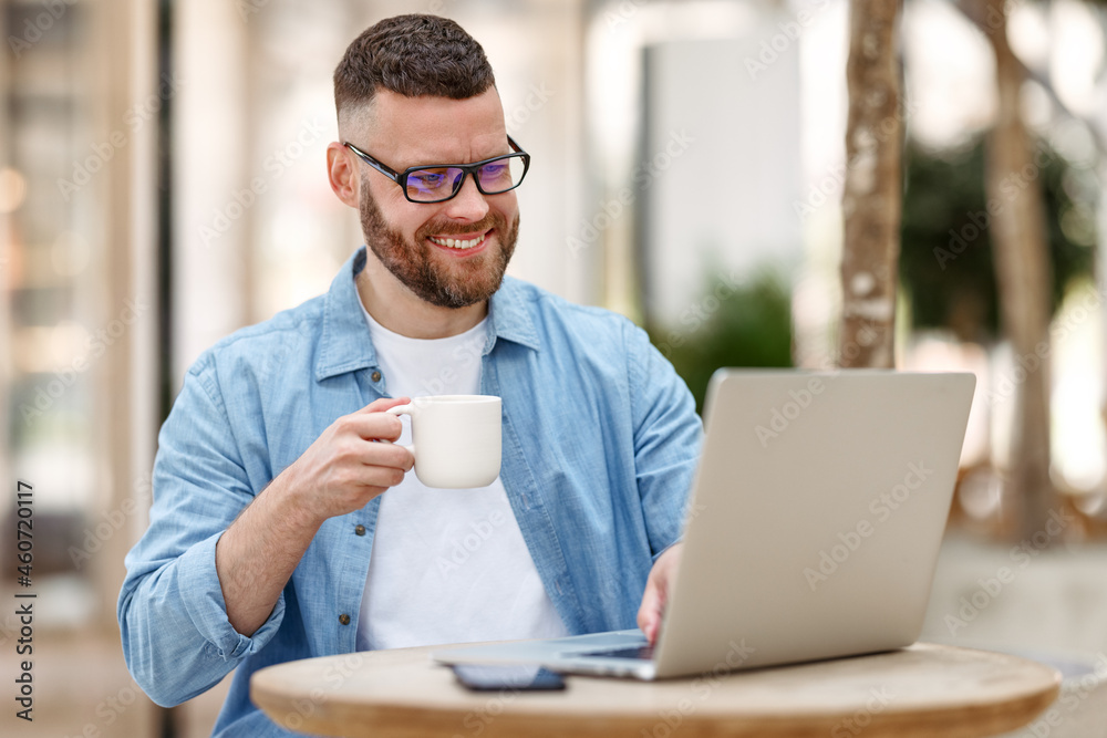 Young happy man freelancer working remotely on laptop, drinking coffee while sitting at outdoor cafe