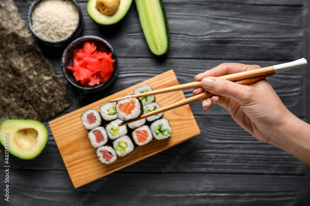Woman taking delicious salmon maki roll with chopsticks on dark wooden background