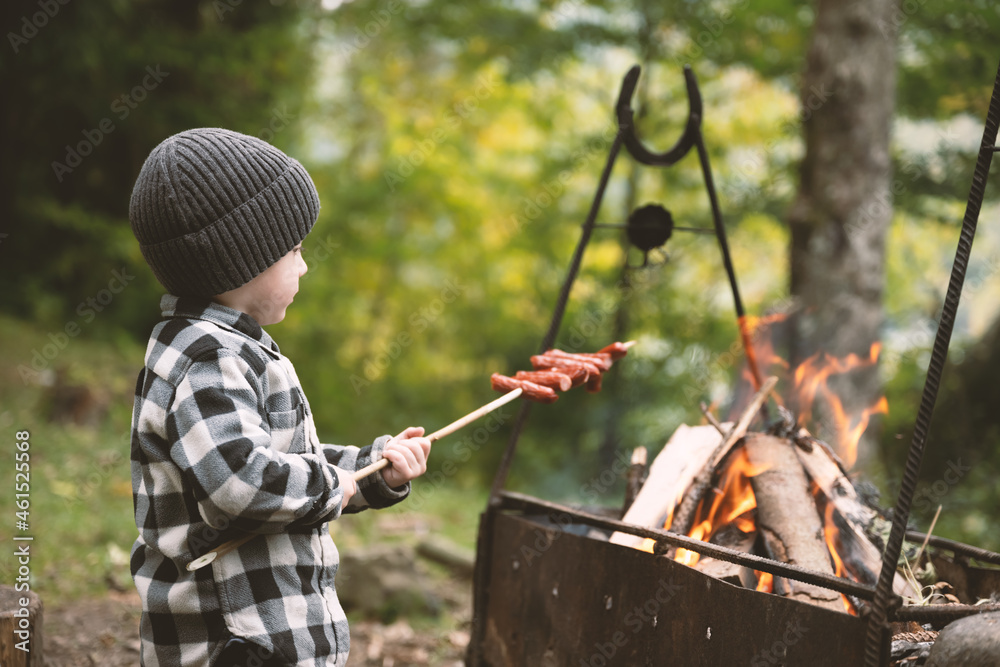 穿着格子衬衫、戴着灰色帽子的小孩在森林的篝火上炸香肠。nat的童年