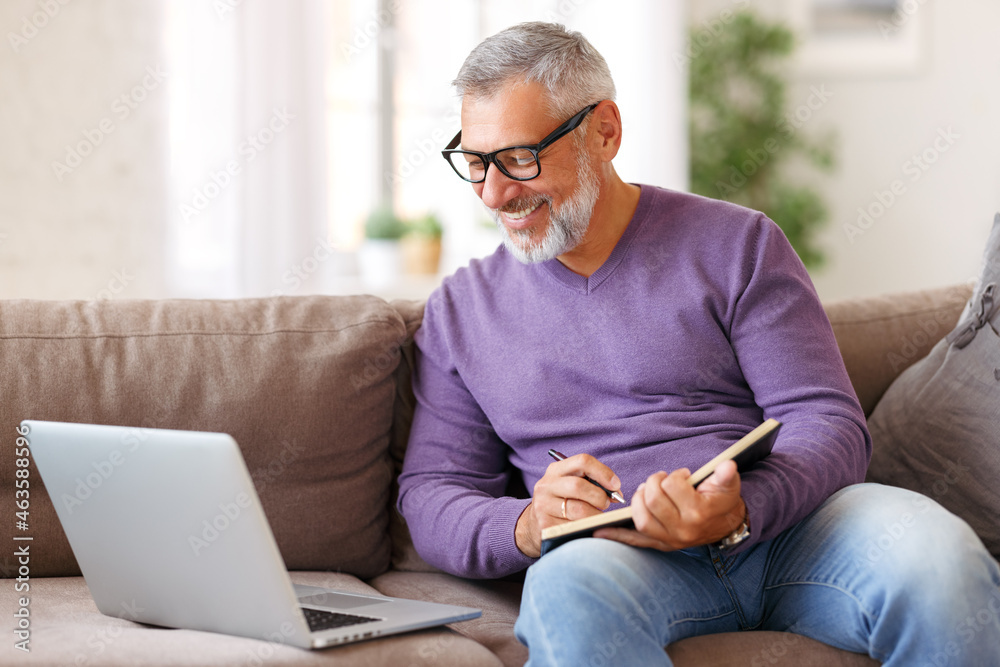 戴着眼镜，坐在沙发上用笔记本电脑远程工作的英俊快乐的老年人