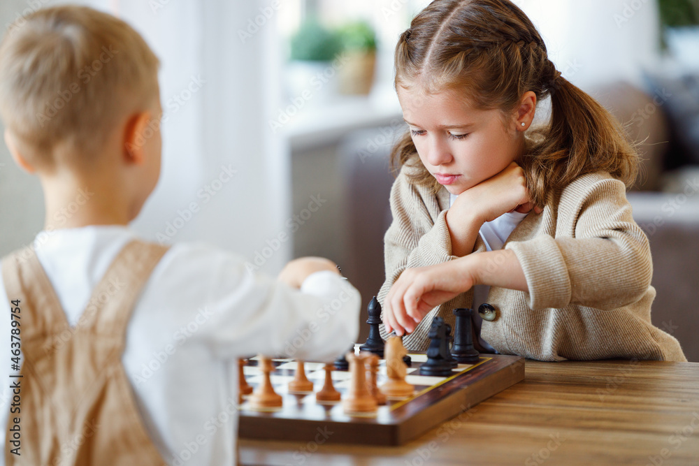 Focused children playing chess game at home while sitting in living room at table with chessboard