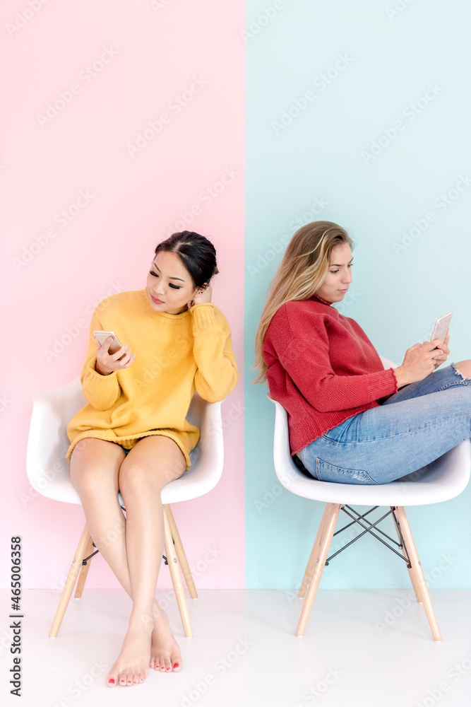 高加索和亚洲女性坐在一起使用手机，背景为粉色和薄荷绿