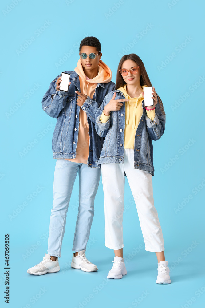 Stylish young couple in hoodies pointing at mobile phones on blue background