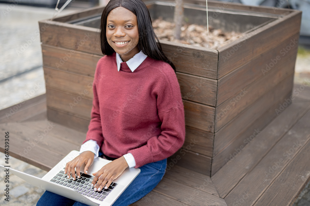 年轻的微笑学生黑人女孩坐在笔记本电脑上打字，看着相机