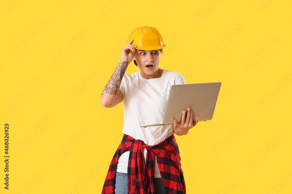 Shocked female construction worker in hardhat using laptop on yellow background