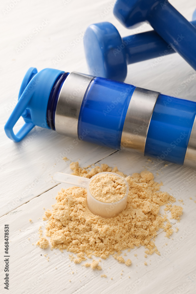 Measuring scoop with powder, bottle with protein shake and dumbbells on white wooden table