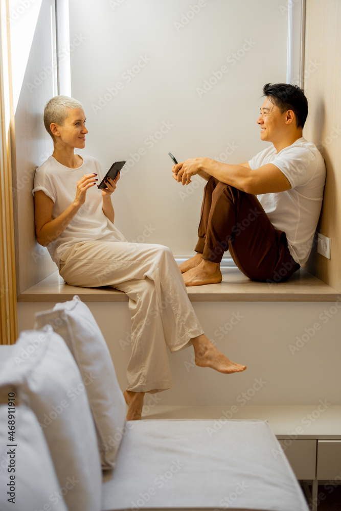 Asian man and caucasian girl look at each other while sit on windowsill and use smartphones at home.